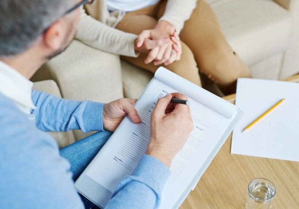 A man writing on paper while sitting next to another person.